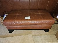 Front view of rectangular brown faux leather ottoman with stitched panels and black wooden legs.