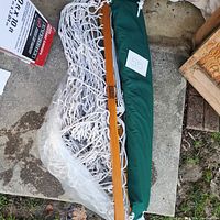 White rope hammock folded on concrete ground with attached green pillow and plastic wrapping. Wooden spreader bars visible.