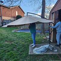 Large tan patio umbrella supported off-center by a curved black metal cantilever stand set on concrete and grass outdoors. Two people handling the umbrella to show scale and condition.