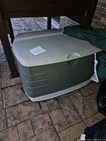Large gray resin plastic outdoor storage box with hinged lid visible, partially covered by a green protective cover on a patio.