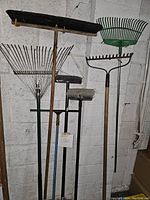 Image showing multiple outdoor tools including rakes with metal and plastic heads, and brooms with worn bristles, standing against a wall.