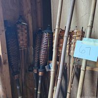 Nine tiki torch holders made of woven bamboo or wood with long bamboo poles leaning against a wall