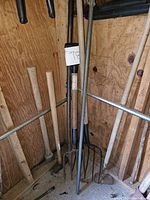 Full view of outdoor tools including pitchforks, axes, and hoes leaning against wall in shed.
