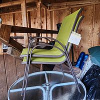 Two stacked metal patio chairs with lime green mesh fabric seats and backs on a round glass table. Wood paneled background.