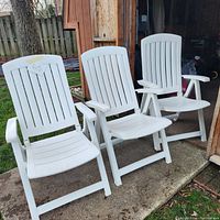 Three white plastic patio chairs arranged outdoors showing their overall condition and design features, including slatted backs, seats, and armrests.