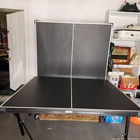 Full view of ping pong table standing upright showing the black playing surface with white borders in a garage setting.