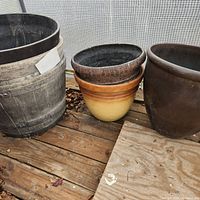 Four large black plastic resin pots and two medium-sized pots shown on a wooden deck, visible dirt and wear signs.