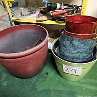 Photo showing six plastic plant pots of various sizes and colors, including a large reddish-brown pot, a green pot, a blue marbled pot, and two smaller red pots stacked inside the green pot.