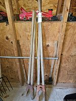 Full view of four garden tools with wooden handles organized against wooden wall including post hole shovels and axe.