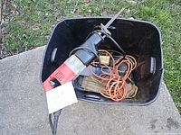 Electric reciprocating saw, several orange power cords, and work gloves in a black plastic bin on a sidewalk next to grass.