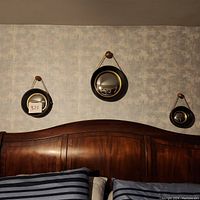 Three convex Witch's Eye mirrors in black ribbed frames with gold inner rims, hanging by gold chains on ornate black and gold hooks above a wooden headboard.