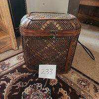 Front angled view of the wicker rattan hinged basket showing its woven pattern, brass accents, and latch front closure. The basket sits on a patterned carpet.