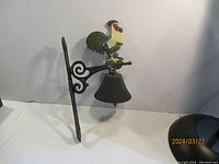Full view of the cast iron rooster bell mounted on a decorative wall bracket against a plain background.