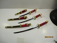Photo shows three ornamental daggers with black sheaths featuring gold-tone fittings, and red cord-wrapped handles lying on a white surface.