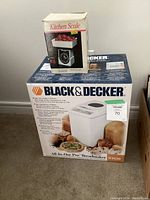 Boxed Black & Decker breadmaker next to boxed kitchen scale on carpeted floor.