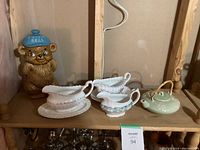 Three ceramic gravy boats and a small teapot displayed on a wooden shelf with a ceramic bear cookie jar in the background.