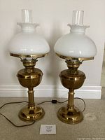 Pair of brass electric hurricane lamps standing side by side on carpet, with white opaque glass shades and clear glass chimneys, showing polished brass bases and bodies.