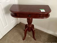 Small dark wood pedestal table shown from the front and angled side view, featuring a single drawer and three curved legs.