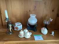 Full view of various small vintage decorative objects placed on wooden shelf, including commemorative ceramic piece, bee salt and pepper and jam set, dog tea light holder, black metal decorative lamp with frosted glass shade and dog figures, glass goblet with floral etching, white egg-shaped vase with floral detail, small dog figurine.