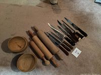 Photo showing 6 wooden bowls, 3 wooden rolling pins, and a set of kitchen knives with wooden handles laid out on a carpeted surface.