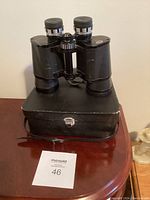 Front view of black vintage binoculars placed on top of the original black carrying case with silver latch, showing overall condition and case style.