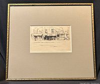 Framed pencil etching showing a street scene with figures and buildings, centered within a beige matte and gold-tone frame.