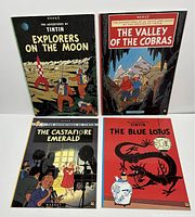 Four vintage Tintin softcover books displayed upright showing front covers: Explorers on the Moon, The Valley of the Cobras, The Castafiore Emerald, and The Blue Lotus. Colors and titles clearly visible.