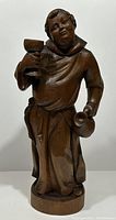 Front view of carved wooden monk holding goblet and jug, showing detailed facial features and robe.