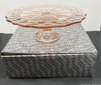 Pink depression glass cake stand displayed on its original box with branded script pattern.