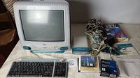 Full view of 1998 Apple iMac G3 desktop with screen, front speakers, floppy disk drive, and power button. Includes keyboard, software disks, manuals, and cables on table.