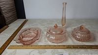 Photo showing set of four pink depression glass pieces: split dish, candy jar with lid, vase, and covered dish.