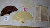Three Chinese folding fans and a letter with Chinese text. Fans include dark brown tortoiseshell-style and two light carved fans, one with pink tassel, displayed on fabric background with ruler for scale.