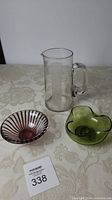 Full view of all three items on a table: large clear glass imperial measure jug with handle, green glass ruffled candle dish, purple swirl patterned dish.