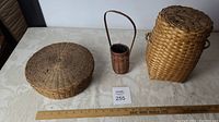 Three woven baskets displayed on a table, including round seagrass basket, a small tubular basket with handle, and a rectangular basket with lid and handles.