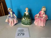 Three Royal Doulton small porcelain figurines (Dinky Do, Belle, Bo Peep) side by side on a surface with auction number card in front.