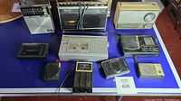 Photo showing 9 vintage radios and 1 cassette recorder laid out on blue cloth surface for condition and model overview.