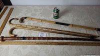 Four walking sticks or canes laid out horizontally on a surface with a soda can for scale, showing two bamboo style canes and two others including a carved dark wood cane with knob handle.