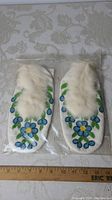 Pair of white moccasins showing overall shape, fur trim, and beadwork design inside clear plastic packaging.
