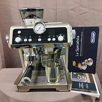 Front view of stainless steel espresso machine with steam wand, portafilter, milk frothing pitcher; original box and manuals in background