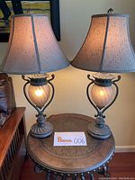 Pair of decorative table lamps lit and placed on a round metal table, showing full height and design details of bases, frosted glass centers, and beige ribbed, paneled shades.