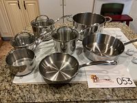 Full set of Farberware Millennium stainless steel cookware arranged on granite countertop with lot number card.