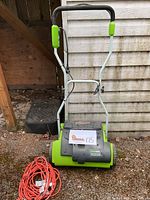 Electric dethatcher unit in green and black, standing upright against a wooden wall with orange extension cord coiled next to it.