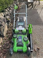 Front angled view showing the Greenworks lawnmower and cordless blower side by side on pavement and near stone wall.