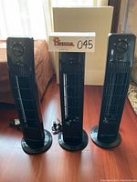 Photo showing three black oscillating tower fans standing on a wood floor in front of a window with partially drawn curtains. Each fan is identical in design and size, with power cords coiled beside or behind the fans.