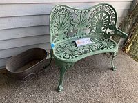 Green painted ornate metal patio loveseat alongside small metal wagon plant holder with wheels