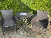 Full photo of patio bistro set showing two brown wicker armchairs with a round folding table with glass top between them, all placed outside on stone ground in front of green hedge.
