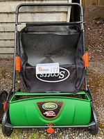 Front view of Scotts Lawn Sweep and Rake showing green body, black frame, orange side knobs, and catch bag with Scotts logo.