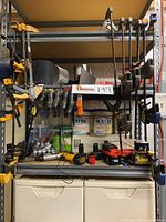 Photo showing shelving with clamps on right side and left side, multiple Irwin clamps, Scotts pole chainsaw attachment on shelf, power tool batteries and cleaning supplies on lower shelf.