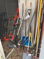 Photo showing assortment of long-handled garden tools including shovels, rakes, and a bucket with tools inside, alongside electric yard equipment against a wall.