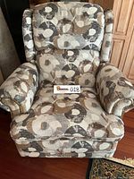 Front view of recliner chair showing patterned fabric upholstery in cream, gray, and brown tones with rivet accents on armrests.